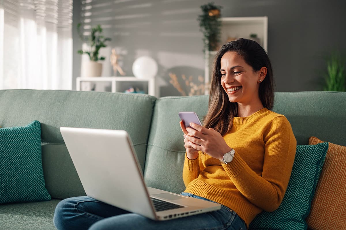 Junge Frau auf Sofa mit Smartphone und Laptop - entspannt erreichbar über Social Media